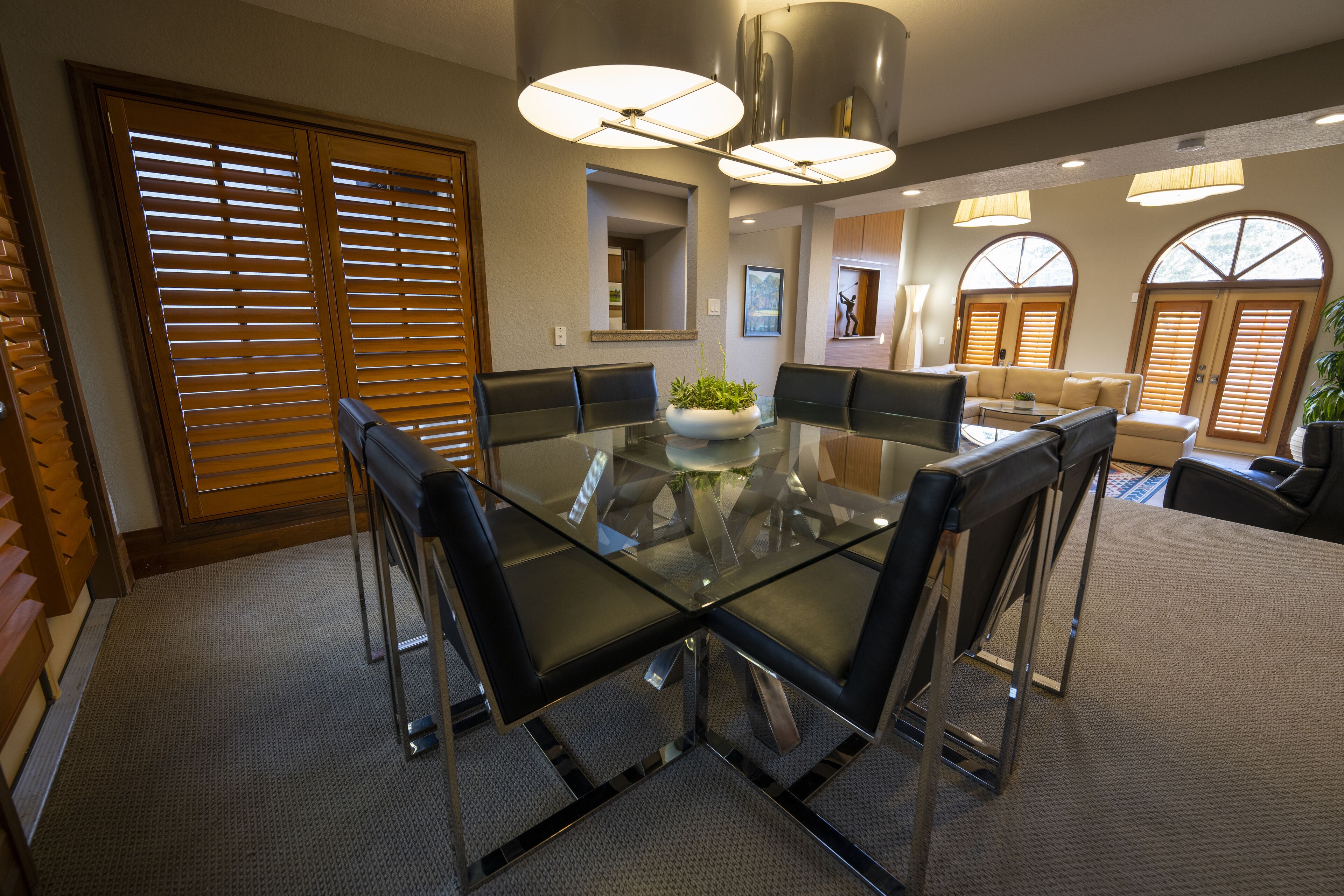 Un comedor moderno con una mesa de vidrio rodeada de sillas, una planta en el centro y un sofá al fondo.