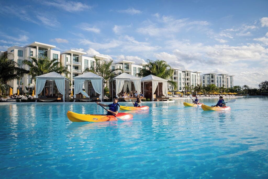 Een zwembad met kayaks, palmbomen en moderne gebouwen op de achtergrond.