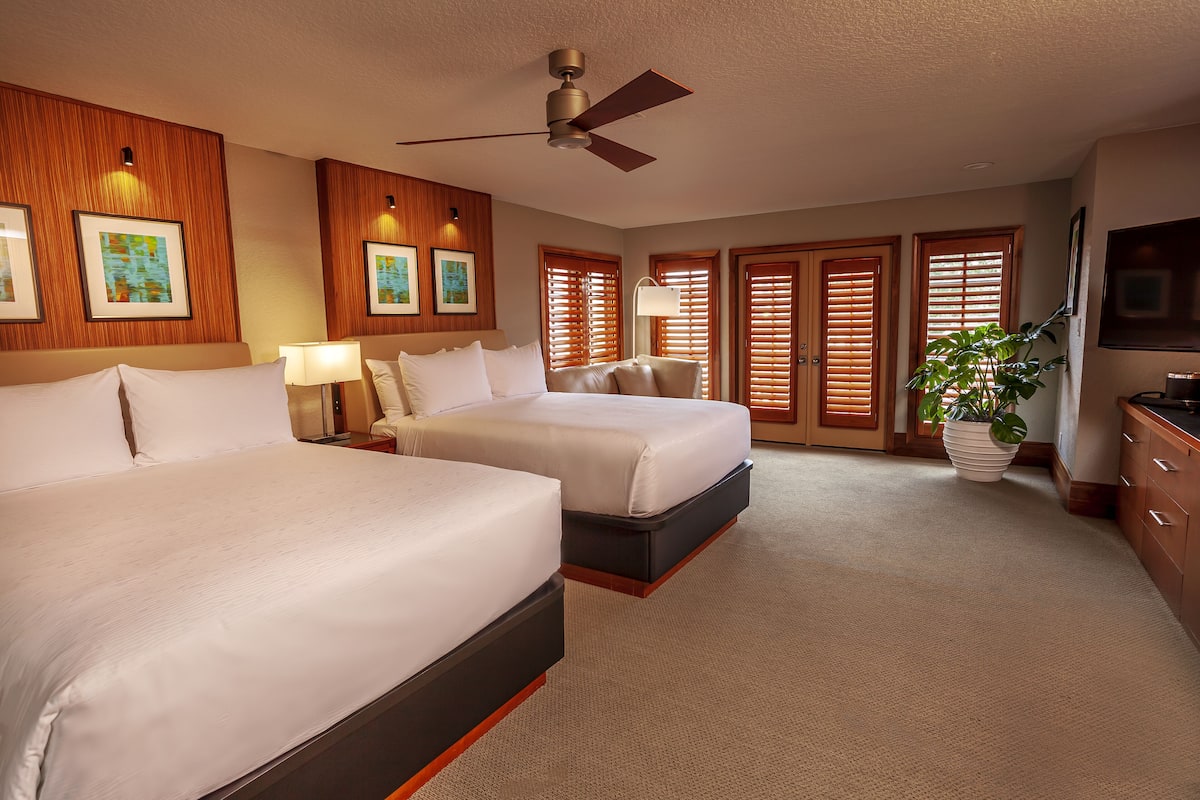 A hotel room with two beds, a ceiling fan, wooden paneling, framed artwork, a potted plant, and a television.