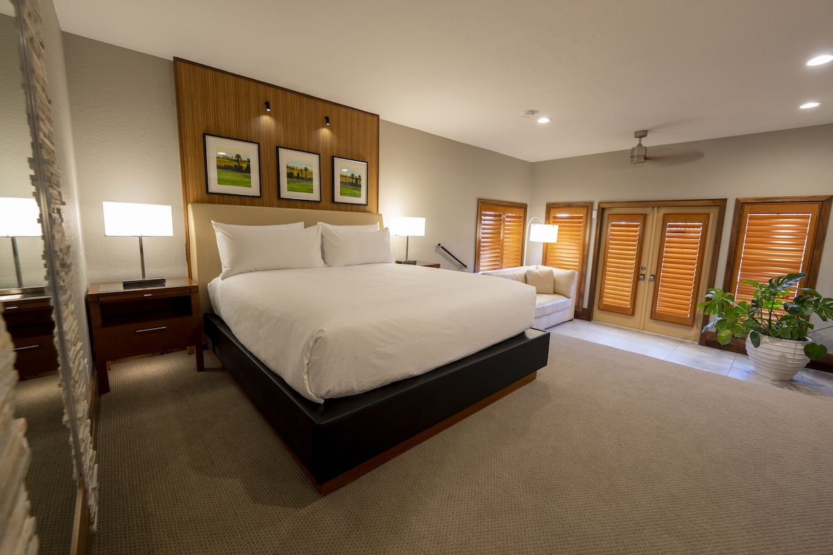 A modern bedroom with a large bed, wooden headboard, bedside tables, and a potted plant.
