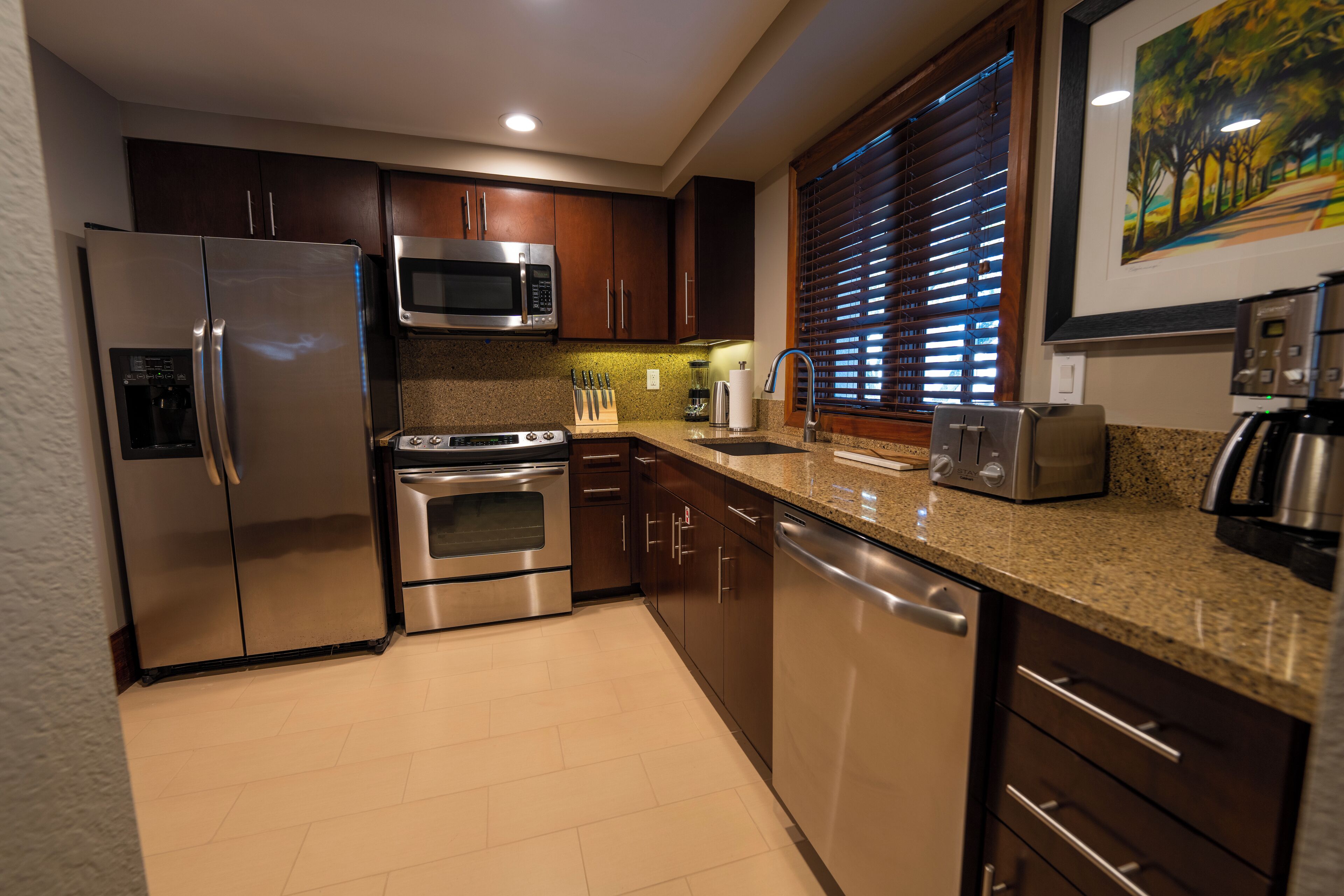 A modern kitchen with stainless steel appliances, wooden cabinets, granite countertops, and a window with blinds.