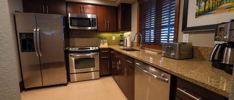A modern kitchen with stainless steel appliances, wooden cabinets, granite countertops, and a window with blinds.