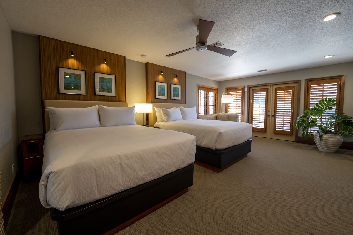 A hotel room with two beds, a ceiling fan, framed artwork on the wall, and a potted plant.