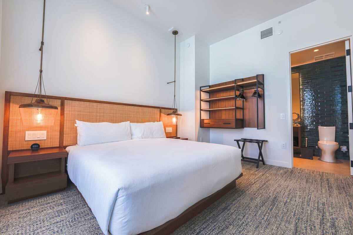 A modern hotel room with a large bed, wooden headboard, bedside tables with lamps, a built-in shelf, and a view of the bathroom.