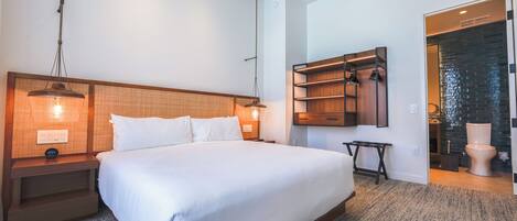 A modern hotel room with a large bed, wooden headboard, bedside tables with lamps, a built-in shelf, and a view of the bathroom.