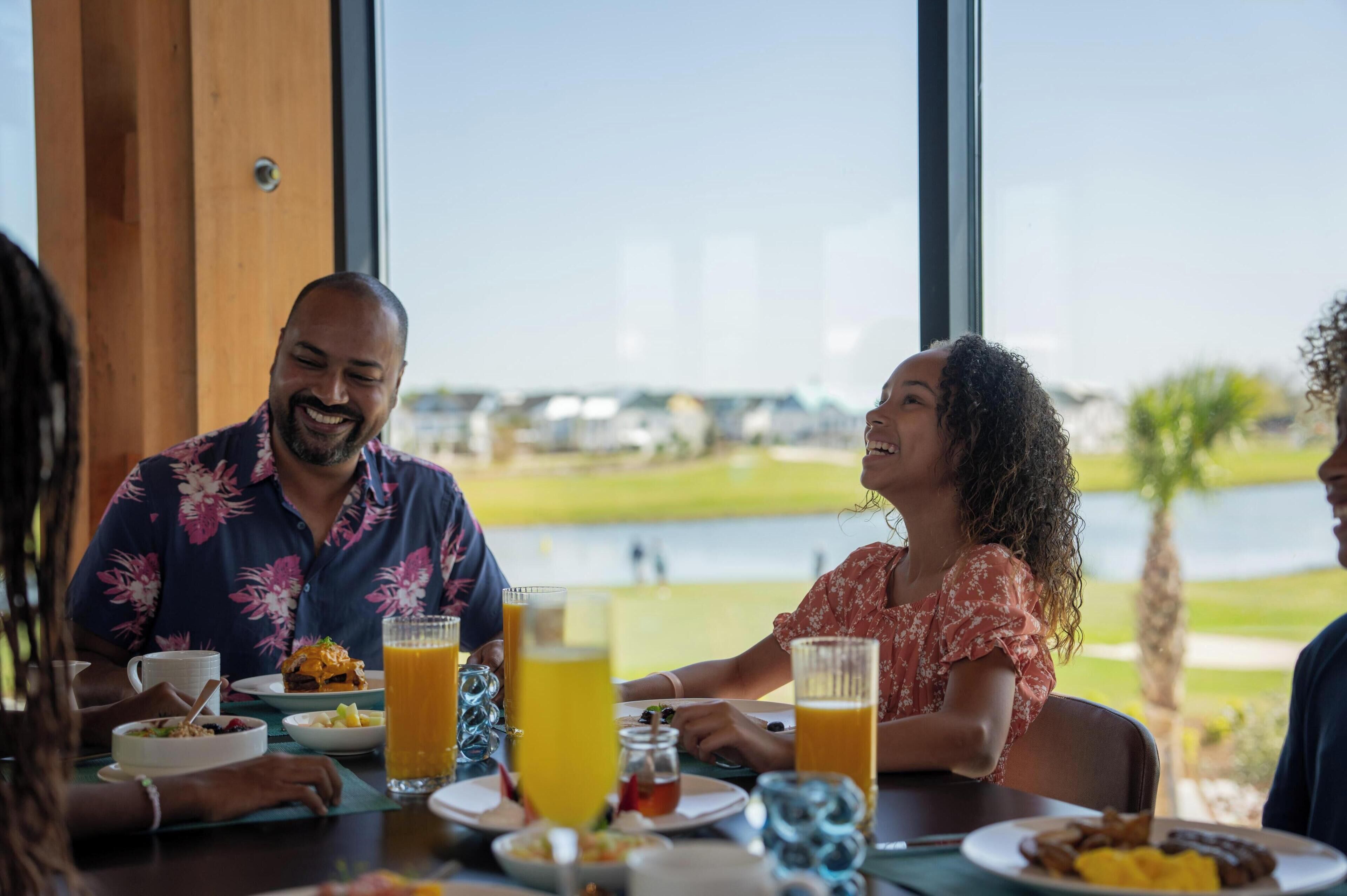 Une table à manger avec des aliments et des boissons pour le petit-déjeuner, de grandes fenêtres donnant sur un terrain de golf et des bâtiments.