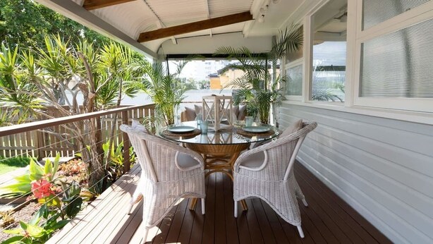 An outdoor patio with a round table and wicker chairs, surrounded by potted plants and a wooden deck. - Boho Beach Lux 250 Meters to Burleigh Sands (Burleigh Heads)