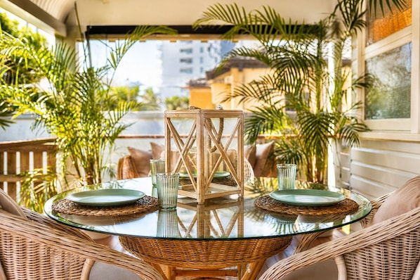 Een glazen tafel met rieten stoelen en een houten lantaarn in het midden, omringd door potplanten.