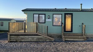 A small, single-story building with a light green exterior, a wooden deck with a ramp, and a door with a decorative glass panel. - Beautiful 2-bed Static Caravan (Newton Stewart)