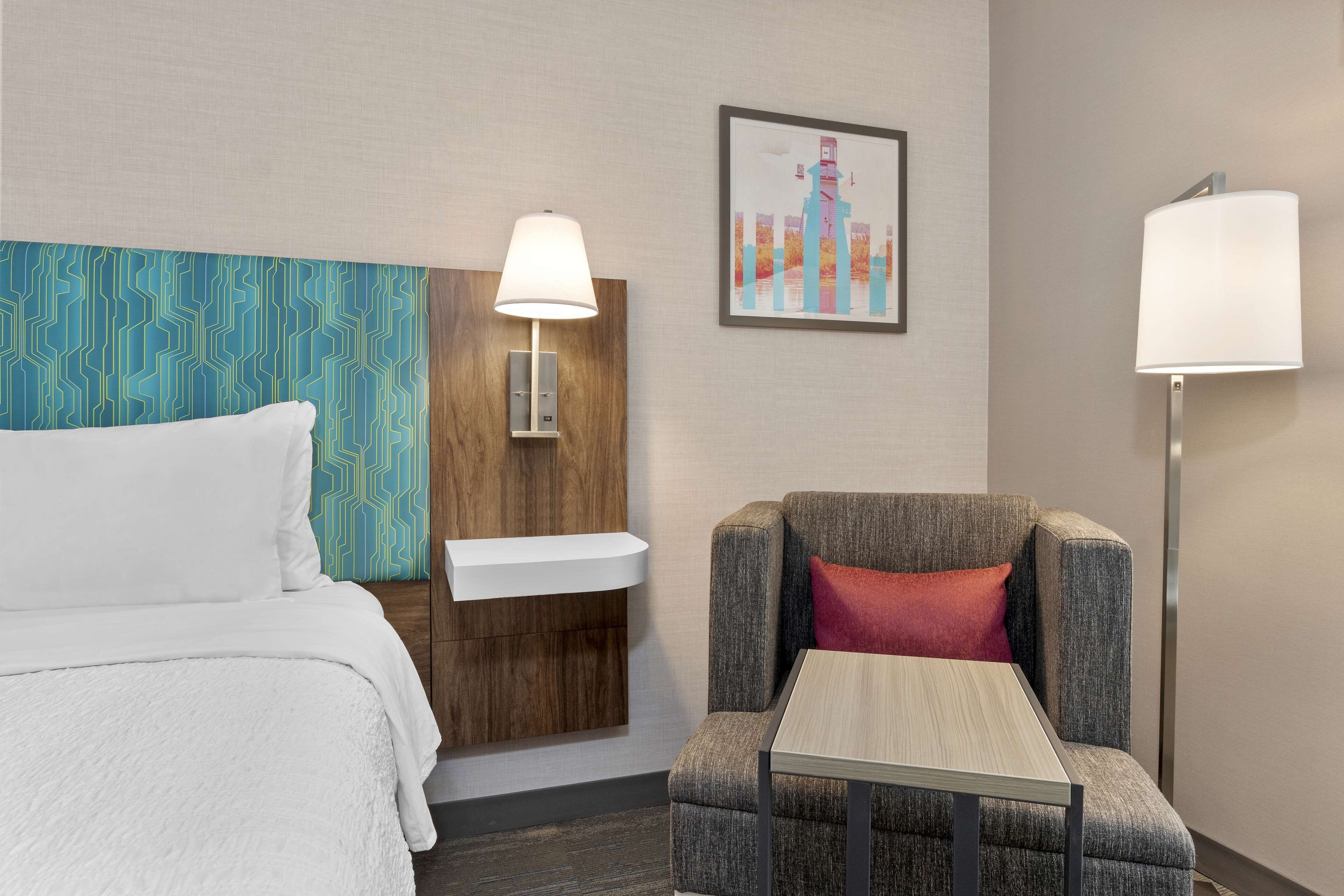 A hotel room with a bed, a chair, a small table, a wall-mounted sink, and two wall lamps.
