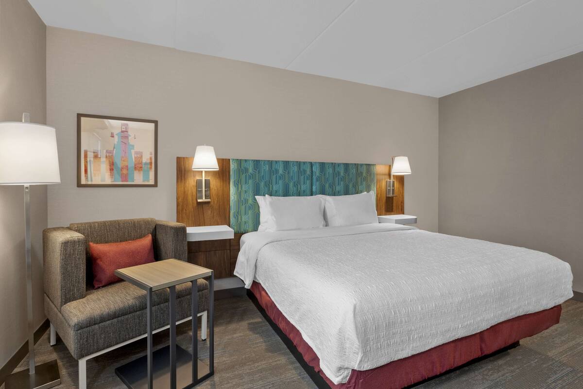 A hotel room with a bed, a sofa, a small table, two bedside lamps, and a wall-mounted TV.