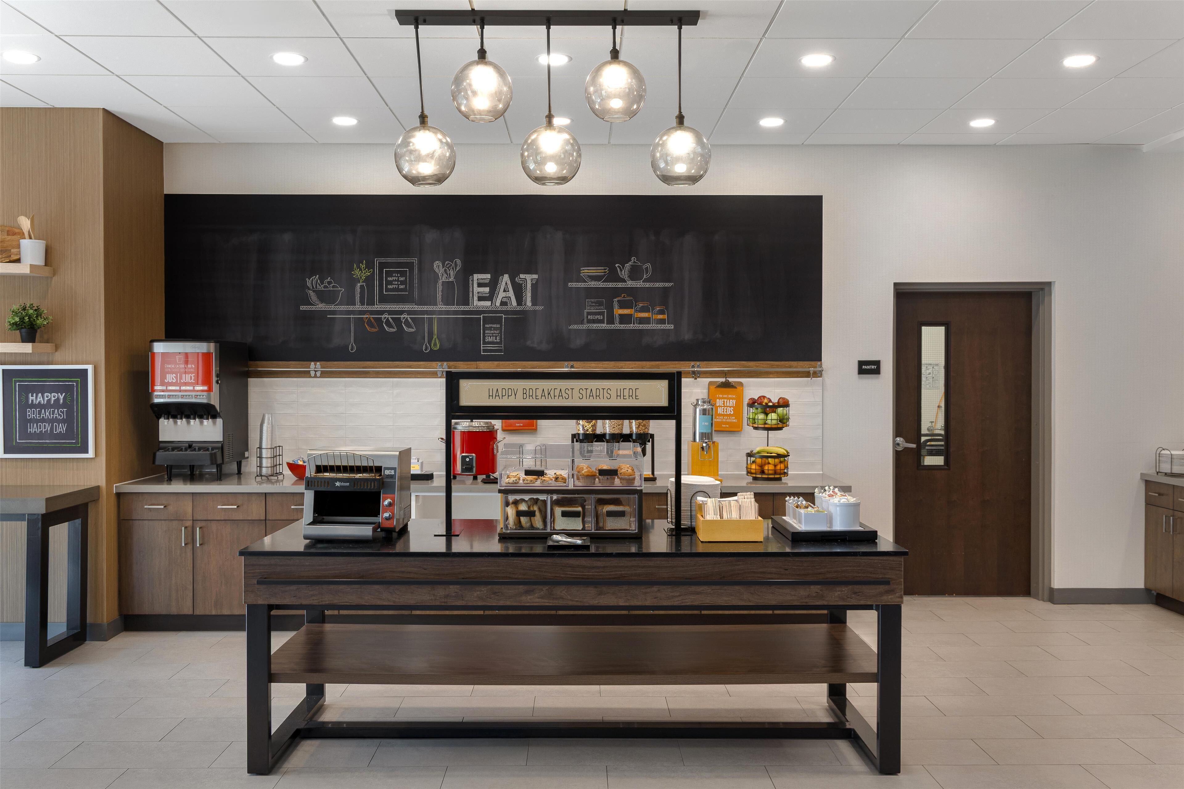 Un espace moderne dédié au petit-déjeuner, avec un tableau noir affichant le menu, des machines à café et une variété d’aliments proposés pour le petit-déjeuner.