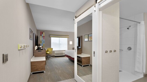 A modern hotel room with a large mirror, a bed, a desk, and a shower area. - Hampton Inn By Hilton Cornwall (Cornwall)