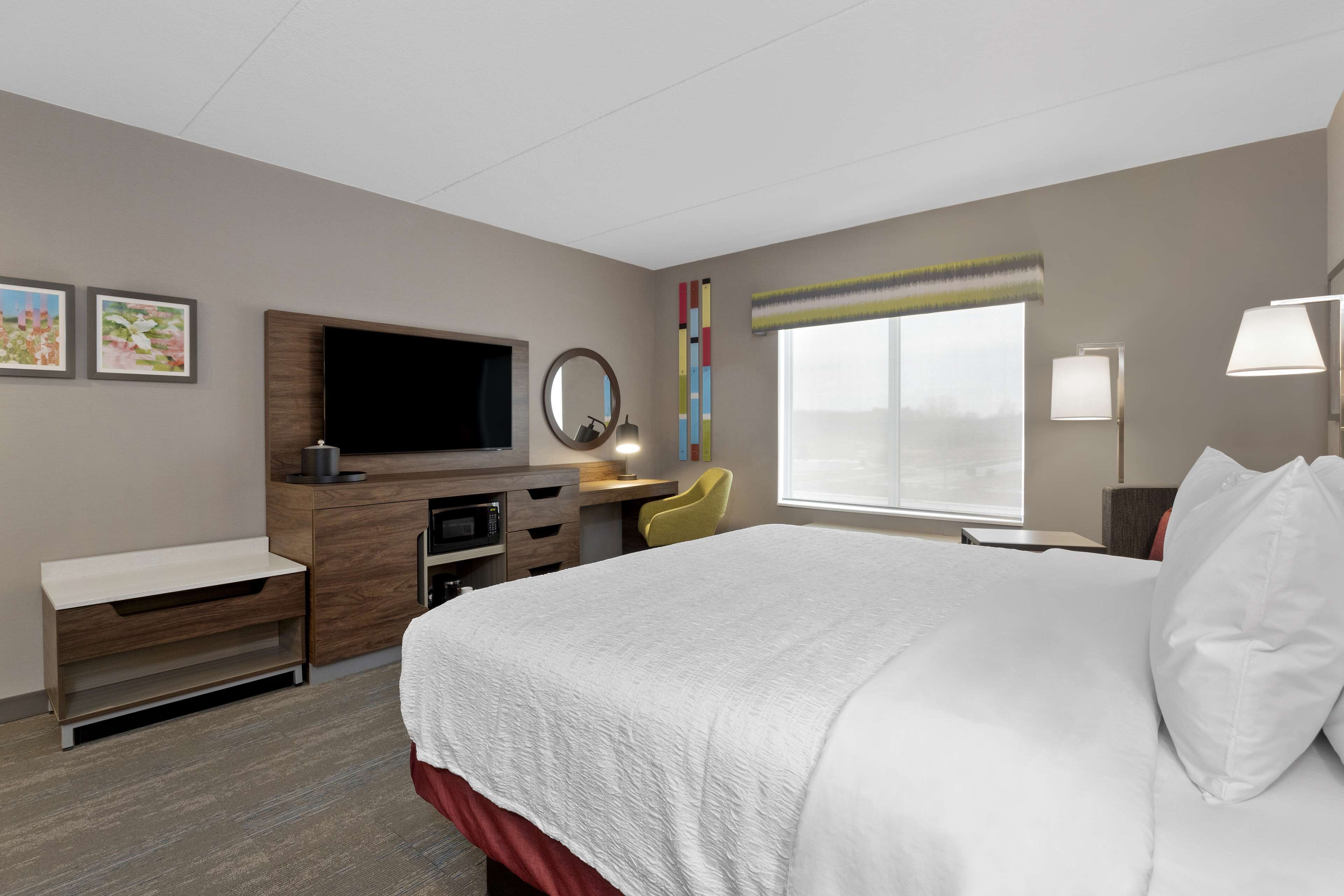 A hotel room with a large bed, a desk with a flat-screen TV, a mirror, a chair, and a window with a view.