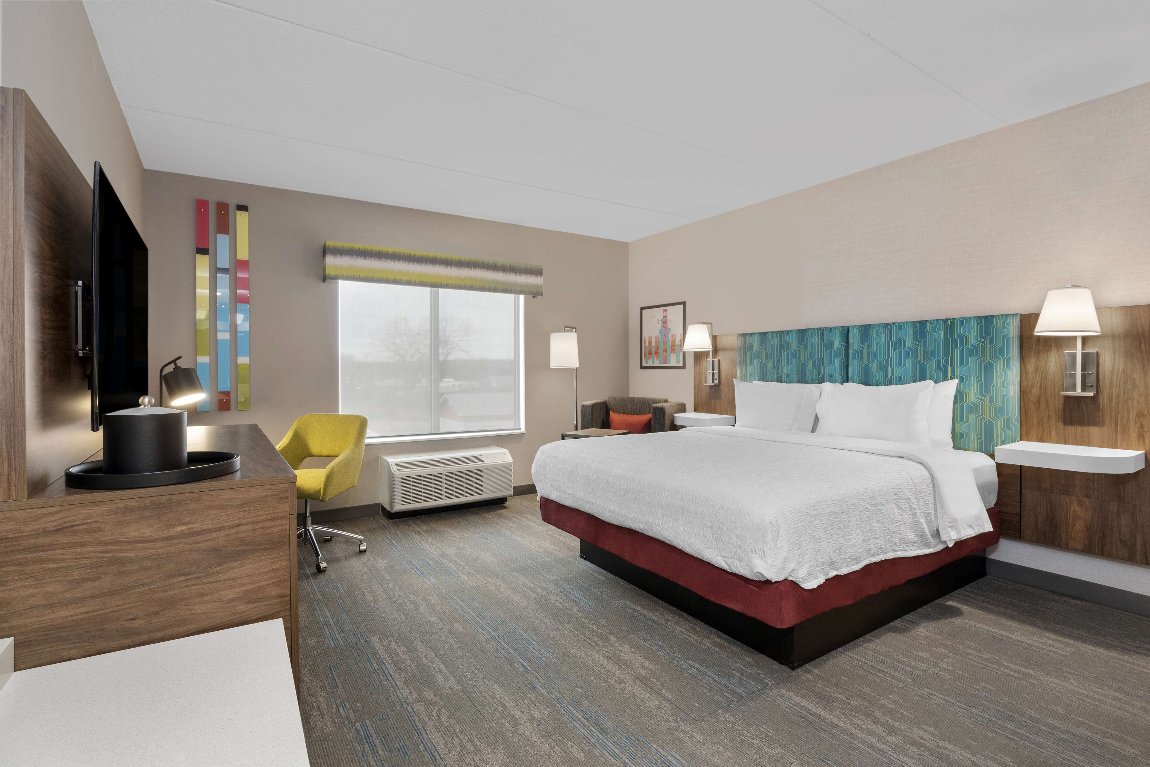 A modern hotel room with a large bed, a desk with a flat-screen TV, a yellow chair, and a window with a view.