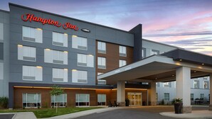 A modern Hampton Inn hotel with a covered entrance, multiple windows, and a landscaped area with a small tree and seating. - Hampton Inn By Hilton Cornwall (Cornwall)