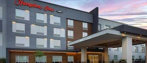 Un moderno hotel Hampton Inn con entrada cubierta, múltiples ventanas y una zona ajardinada con un pequeño árbol y asientos.
