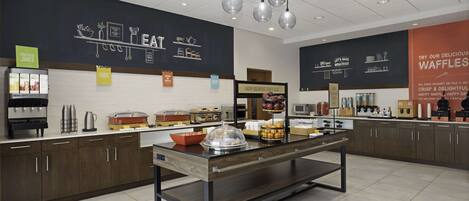 Zona de desayuno tipo buffet con distintas estaciones de comida, incluyendo un cartel que dice 'Aquí comienza un desayuno feliz' y una presentación de gofres.