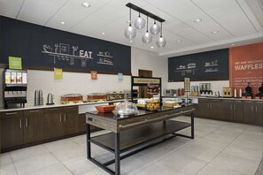 A breakfast buffet area with various food stations, including a sign for 'Happy Breakfast Starts Here' and a display for waffles.
