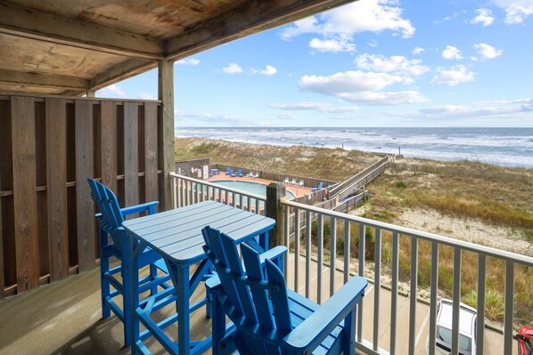 A covered outdoor seating area with blue chairs and a table, overlooking a beach with a wooden deck and lounge chairs.