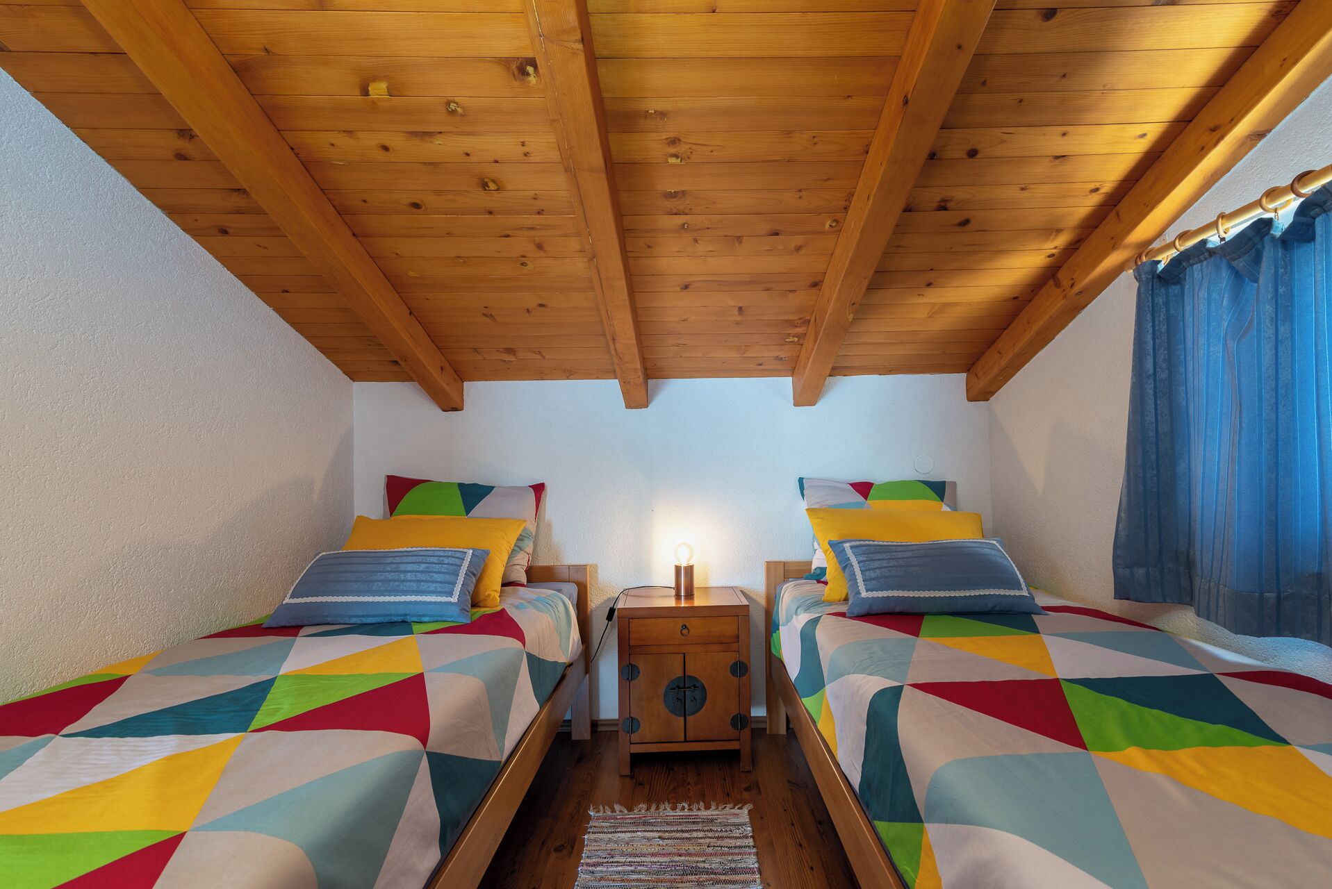 A room with two beds, wooden ceiling beams, a nightstand, and a window with curtains.