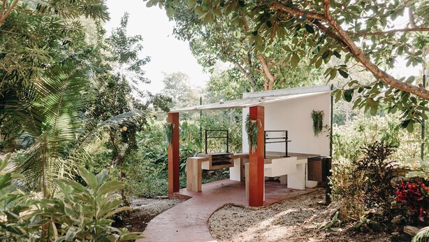 Un espace extérieur aménagé, abrité et doté d’un banc et d’une table, entouré d’une végétation luxuriante et d’arbres.