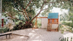 Une structure de jeu en bois, surmontée d’un toit bleu, entourée d’un feuillage dense et posée sur un sol sablonneux.