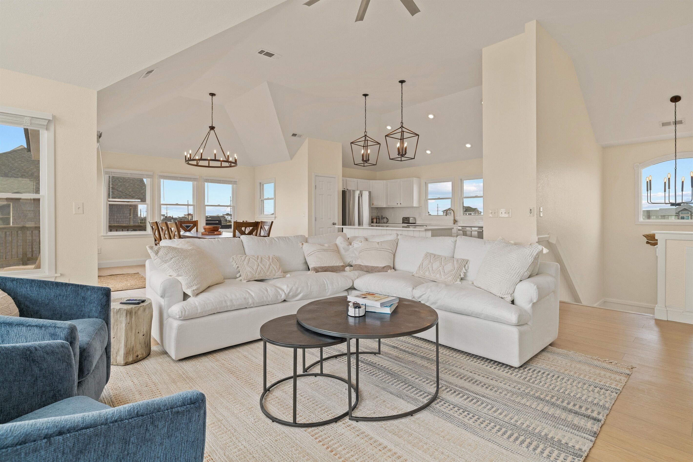 A spacious living room with a large L-shaped white sectional sofa, a round coffee table, and a blue armchair. There are two pendant lights above the coffee table and a chandelier in the dining area.