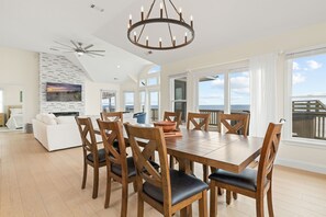 Uma ampla sala de jantar com mesa e cadeiras de madeira, um lustre moderno e grandes janelas com vista para o oceano.
