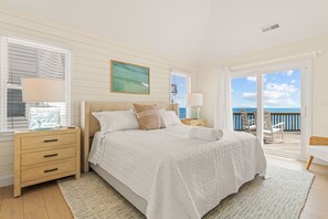 Quarto com cama grande, mesa de cabeceira, luminária, pintura e vista para o oceano.