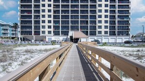 Un hotel a più piani con un ingresso centrale, una passerella in legno che conduce all'ingresso e una spiaggia sabbiosa in primo piano.
