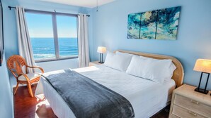 Una camera da letto con un grande letto, vista sull'oceano, una poltrona in vimini e un comodino con una lampada.