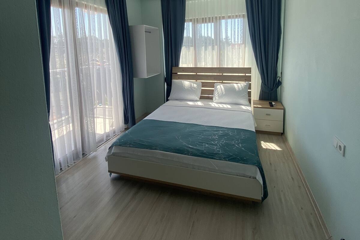 Un dormitorio con cama, mesita de noche, ventilador de techo y ventana con cortinas.