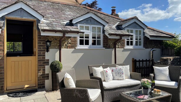 A cozy outdoor seating area with wicker furniture and a glass table, adjacent to a house with a sloped roof and a chimney. - The Coach House at Lackington Farm (Dorchester)