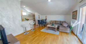 A modern living room with a sofa, coffee table, and two bar stools. There is a kitchen area with a window and a dining table.