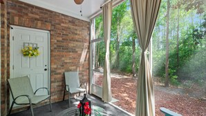 Una habitación con pared de ladrillo, una puerta blanca adornada con un ramo de flores, dos sillas, una mesa con un jarrón y un ventanal con cortinas que da a un bosque.