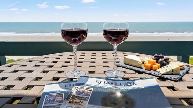 Dos copas de vino tinto, una tabla de quesos y un menú sobre una mesa con vista al océano.