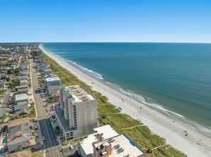 An aerial view of a coastal city with a long beachfront, high-rise buildings, and a clear blue sky.