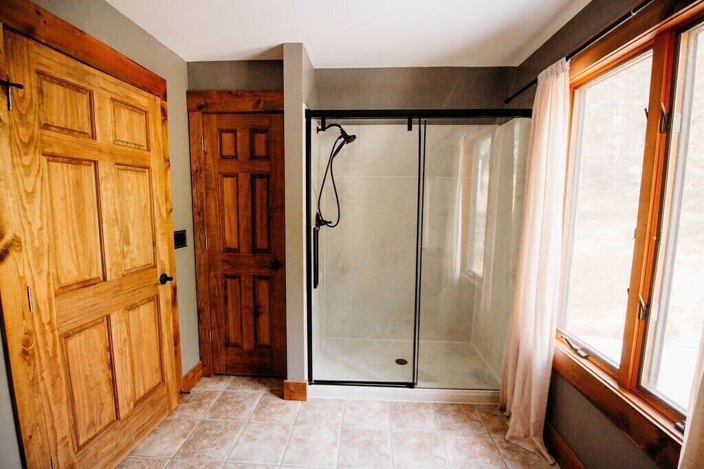 Una habitación con puerta de madera, cabina de ducha de vidrio y ventana con cortinas.