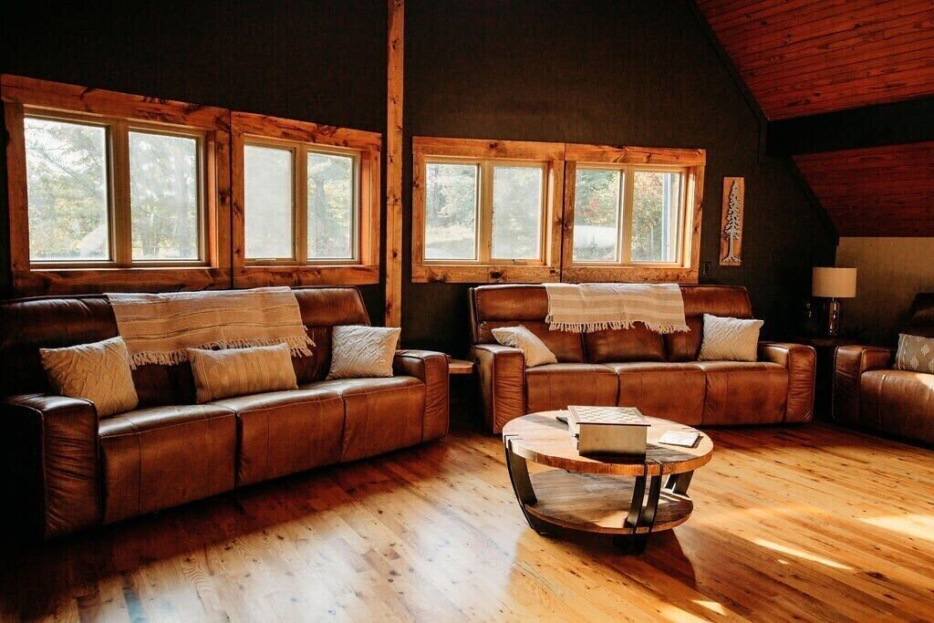 A cozy living room with two leather sofas, a round coffee table, and large windows with wooden frames.