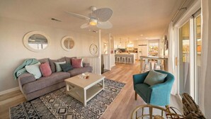 A modern living room with a sofa, coffee table, and armchair, a dining area with bar stools, and a kitchen in the background. - Cozy Home w/ 2 Pools, Vball Court, & Ocean Views (Galveston)