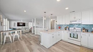 A modern kitchen with white cabinets, a central island, and a dining area with a table and chairs. - Cozy Home w/ 2 Pools, Vball Court, & Ocean Views (Galveston)