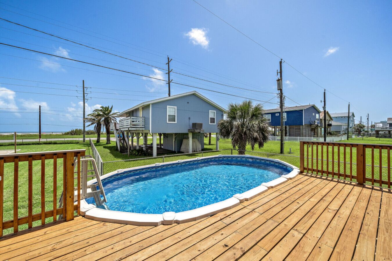Une terrasse en bois avec une piscine, une maison dotée d’une terrasse couverte et un ciel d’un bleu éclatant.