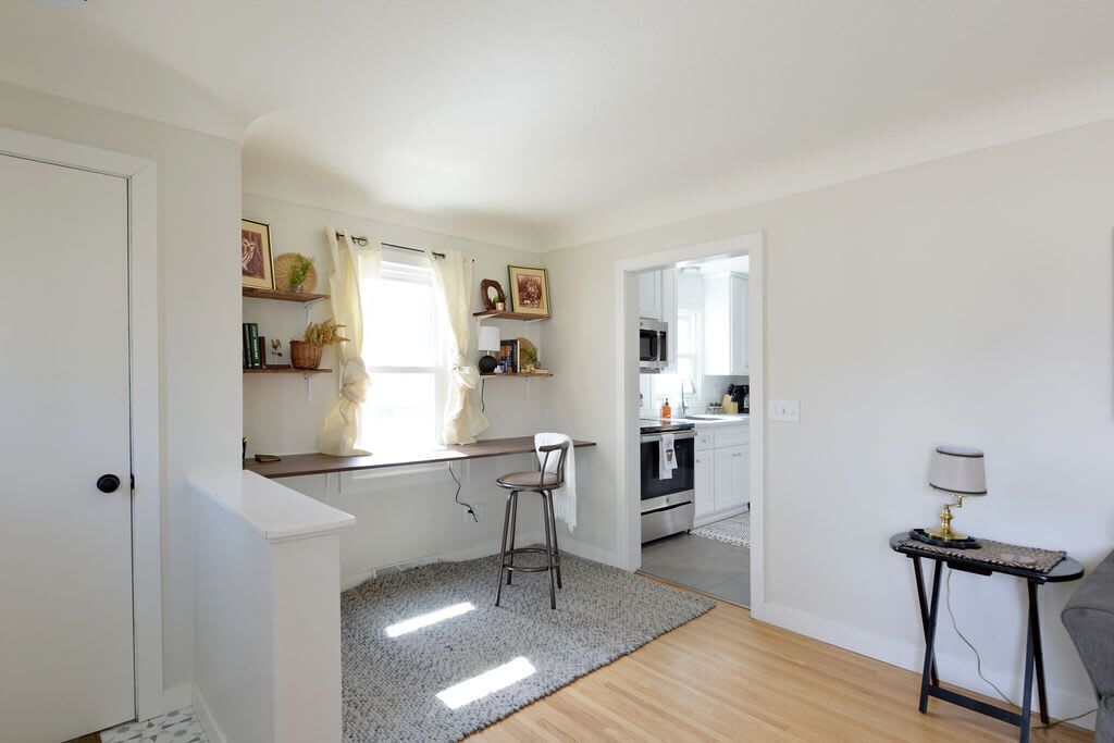 A modern kitchen with white cabinets, a wooden countertop, and a bar stool. There is a window with curtains, a small table with a lamp, and a grey rug on the floor.