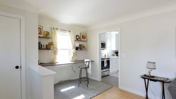A modern kitchen with white cabinets, a wooden countertop, and a bar stool. There is a window with curtains, a small table with a lamp, and a grey rug on the floor.