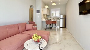 A modern living room with a pink sofa, a marble coffee table with fruit, and a kitchen area with a fridge and bar stools. - Nissaki Anemone Luxury Apartment with Jacuzzi (Nissaki)
