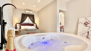 A hotel room with a large hot tub, a bed with curtains, a bedside table, and a painting on the wall. - Nissaki Anemone Luxury Apartment with Jacuzzi (Nissaki)