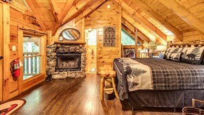 A log cabin bedroom with a stone fireplace, wooden ceiling beams, and a large bed with a plaid bedspread. - Romantic Creekside Cabin-Secluded Mountain Escape (Pigeon Forge)