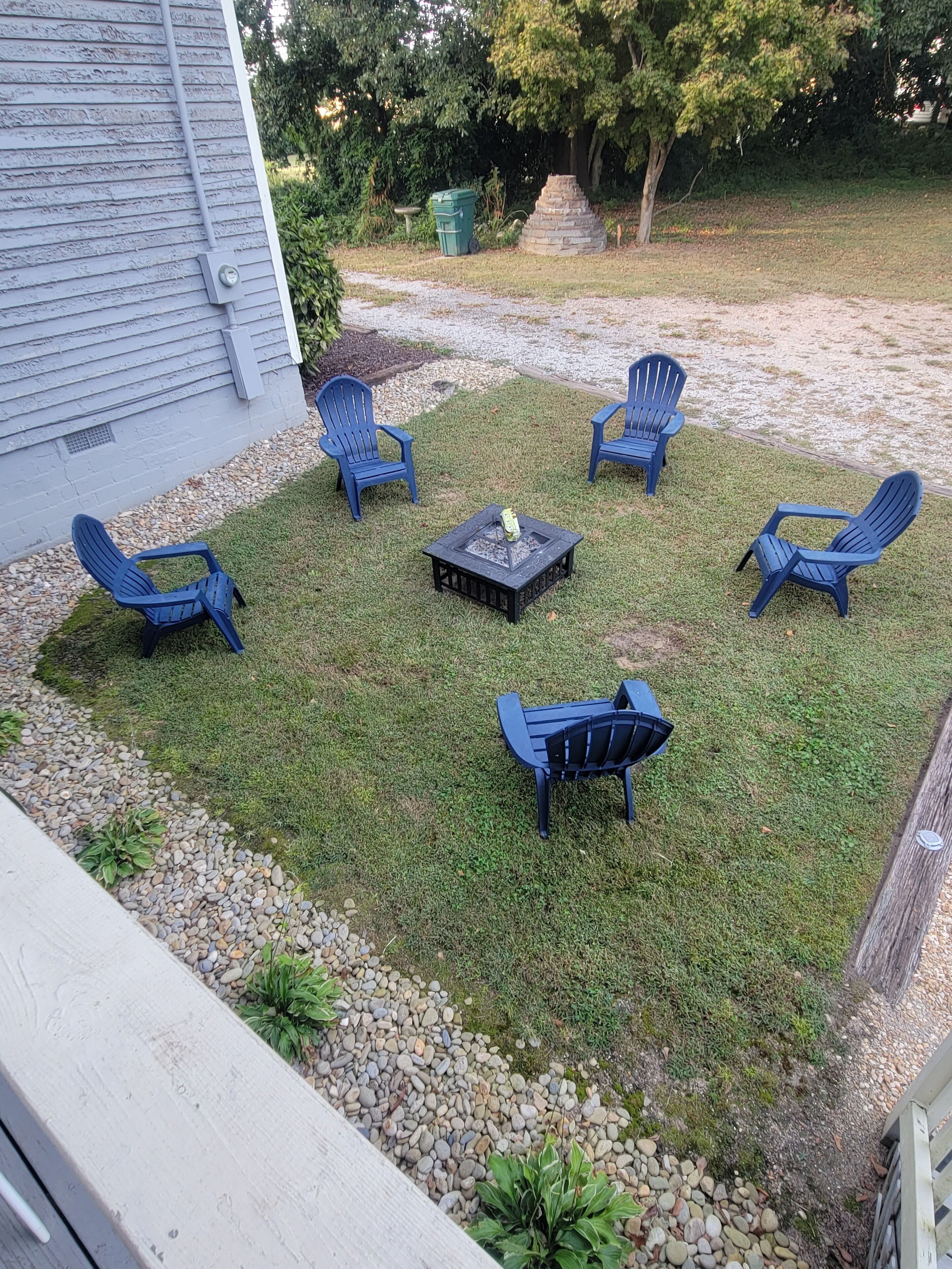 A backyard with five blue Adirondack chairs arranged around a square table, a stone structure, and a green trash bin.
