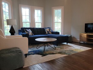 Una sala de estar con un sofá azul, una mesa de centro redonda, una lámpara y un televisor sobre un mueble de madera.